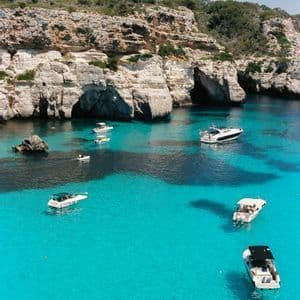 Vista aerea di barche in acque turchesi limpide in una cala circondata da scogliere rocciose.