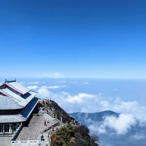 Veduta dall'alto di un edificio tradizionale in cima a una montagna, con persone che percorrono un sentiero affacciato su un mare di nuvole e un vasto cielo blu.