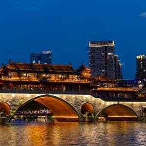 Un ponte tradizionale cinese illuminato con edifici in cima attraversa un fiume di notte, con moderni grattacieli sullo sfondo sotto una luna crescente.