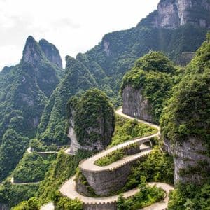 Una strada tortuosa con molti tornanti e muretti a secco si inerpica su una ripida e verde catena montuosa sotto un cielo nuvoloso.