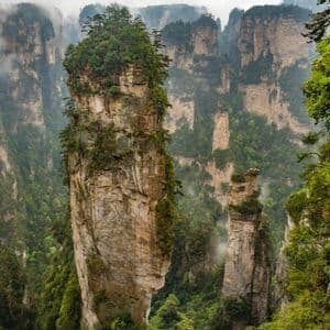 Alte formazioni rocciose a pilastro, ricoperte di vegetazione verde, si ergono da una fitta foresta, mentre la nebbia si insinua tra le valli.