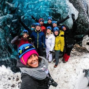 Un voyage de groupe WeRoad portant des casques et des vestes d'hiver sourit pour un selfie à l'intérieur d'une grotte de glace bleue translucide.