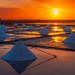 Pyramidenförmige Haufen weißen Salzes liegen in flachen Verdunstungsbecken und spiegeln einen leuchtend orangefarbenen Sonnenuntergang wider.