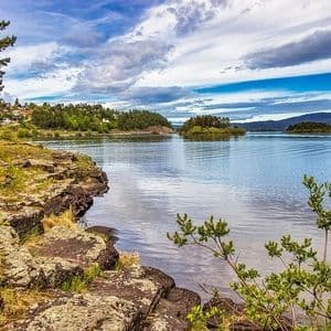 Una costa rocciosa incontra un lago calmo con diverse piccole isole alberate e colline lontane sotto un cielo parzialmente nuvoloso.