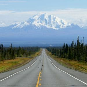 A straight paved road stretches through a forest of evergreens towards a large, snow-capped mountain range in the distance.