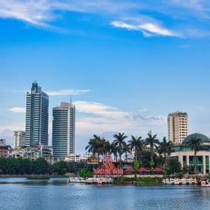 Uno skyline cittadino con alti edifici e alberi verdi, visto attraverso uno specchio d'acqua con pedalò a forma di cigno ormeggiati lungo la riva.