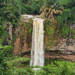 Una potente cascata scende da una scogliera rocciosa muschiosa, circondata da una fitta foresta tropicale sotto un cielo parzialmente nuvoloso.