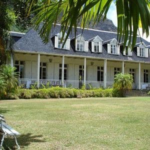 Una casa bianca in stile coloniale con tetto scuro e veranda si erge dietro un ampio prato verde, incorniciata da lussureggianti palme.