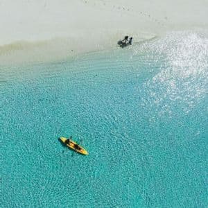Vista aerea di due kayak gialli in acque limpide e turchesi accanto a una spiaggia di sabbia bianca con persone e pedalò.