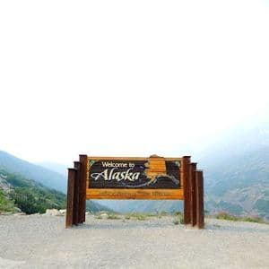 A wooden 'Welcome to Alaska' sign stands on a gravel patch overlooking a winding mountain road and hazy peaks.