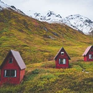 Four small red A-frame cabins are scattered on a grassy hillside at the base of a large, snow-capped mountain under a cloudy sky.