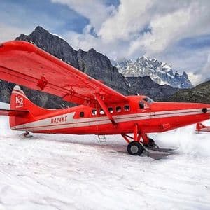 A red propeller plane fitted with skis is parked on a snowy glacier, with a rugged mountain range in the background.