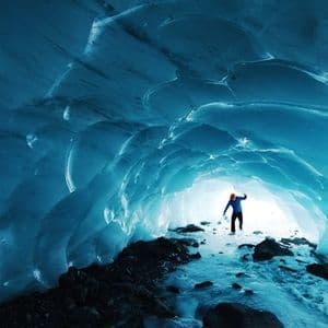 A person in a blue jacket and red hat stands inside a glowing blue ice cave, looking towards the bright entrance.