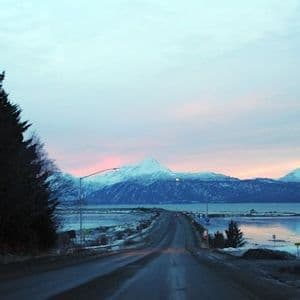 A wet road runs alongside a calm body of water, leading towards snow-capped mountains under a pastel twilight sky.