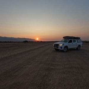 Un pick-up bianco con tenda da tetto è parcheggiato su una strada sterrata in un vasto paesaggio desertico mentre il sole tramonta dietro le montagne.