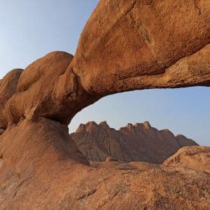 Un imponente arco naturale di formazioni rocciose rossastre, con vista su montagne lontane attraverso l'apertura sotto un cielo sereno.
