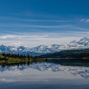 A snow-capped mountain range and a green treeline are perfectly reflected in the calm waters of a lake under a blue sky.