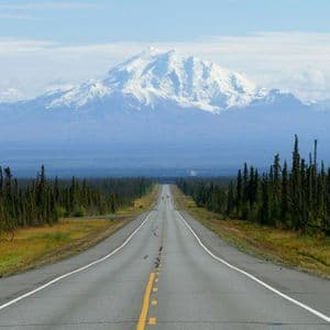 A straight paved road stretches through a dense evergreen forest towards a large, snow-covered mountain range under a partly cloudy sky.