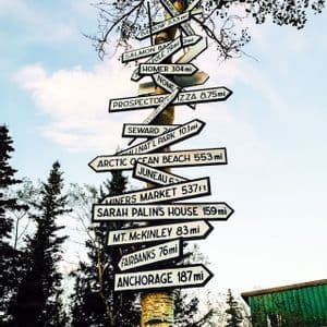 A birch tree trunk covered in directional signs pointing to various Alaskan locations like Anchorage, Fairbanks, and Mt. McKinley.