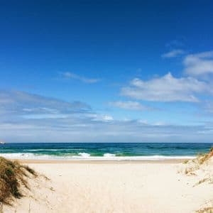 Un sentiero sabbioso tra due dune erbose conduce a una spiaggia di sabbia bianca con onde blu dell'oceano sotto un cielo parzialmente nuvoloso.