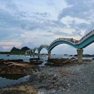 Eine hellblaue, mehrbogige Brücke erstreckt sich vom felsigen Ufer über das Meer zu einer kleinen Insel unter einem bewölkten Himmel.