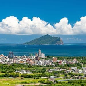 Eine Küstenstadt, umgeben von Grün, blickt auf eine große Insel im Meer unter blauem Himmel mit bauschigen weißen Wolken.