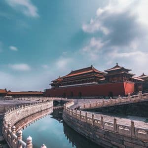 Ein gewundener Wassergraben mit steinerner Balustrade spiegelt einen traditionellen roten chinesischen Palast unter einem gestreiften blauen Himmel wider.