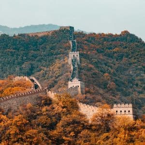 Eine lange Steinmauer mit Wachtürmen zieht sich einen steilen Berg hinauf und hinunter, der von dichten, herbstlich gefärbten Bäumen bedeckt ist.