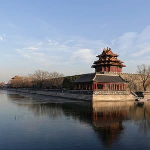 Ein chinesischer Eckturm auf einer Steinmauer spiegelt sich im stillen Wasser eines Wassergrabens unter klarem Himmel.