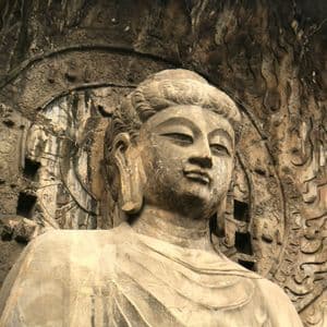 Una grande statua di Buddha in pietra, erosa dal tempo, scolpita in una parete rocciosa e circondata da elaborati bassorilievi.