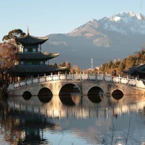 Un ponte in pietra ad arco e una pagoda che si riflettono in un lago calmo, con montagne innevate visibili in lontananza.