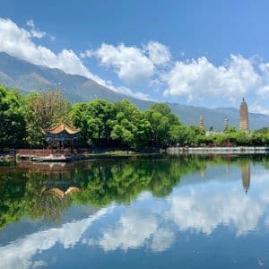 Un padiglione tradizionale e tre pagode si affacciano su un lago, riflettendo gli alberi verdi, le montagne distanti e il cielo azzurro e nuvoloso.