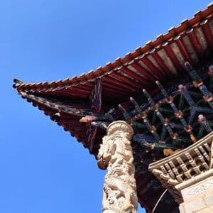 Froschperspektive auf eine kunstvoll verzierte Steinsäule mit Drachenschnitzereien, die das rot-gedeckte Dach eines traditionellen Tempels vor blauem Himmel trägt.