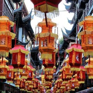 Reihen von roten und orangen chinesischen Laternen mit gelben Quasten hängen über einer Straße zwischen traditionellen Gebäuden.