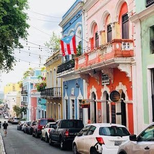 A Puerto Rican flag hangs from a balcony on a street lined with colorful buildings and parked cars.
