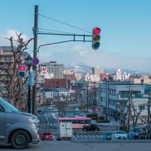 Una vista ampia di una strada cittadina giapponese con traffico, edifici e montagne innevate distanti sotto un cielo azzurro chiaro.