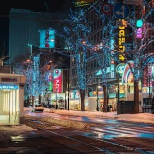 Una strada cittadina innevata di notte illuminata da insegne al neon colorate e alberi avvolti da luci blu, con binari del tram in primo piano.