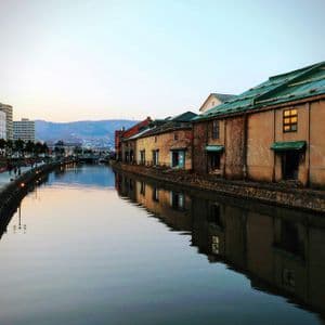 Vecchi magazzini con tetti verdi si riflettono in un tranquillo canale cittadino al crepuscolo, di fronte a una passeggiata illuminata dove le persone camminano.