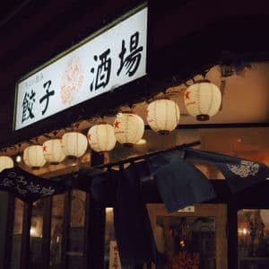 Lanterne di carta bianche e illuminate sono appese in fila sotto un'insegna luminosa con caratteri giapponesi, a indicare un ingresso di notte.