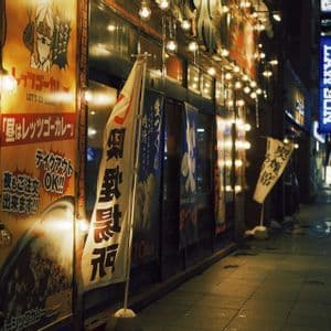 Una vetrina illuminata con insegne e bandiere giapponesi si affaccia su una strada cittadina bagnata di notte, con un'insegna blu luminosa visibile in lontananza.