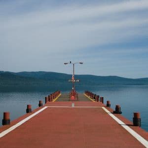 Un lungo molo rosso si estende su un lago tranquillo verso lontane montagne boscose sotto un cielo blu.