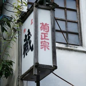 Insegna giapponese verticale con caratteri neri e rossi in una cornice di legno, fissata alla parete bianca di un edificio accanto a una finestra.