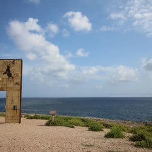 Una grande installazione artistica, erosa dalle intemperie e a forma di portale, si erge su una costa ghiaiosa che si affaccia sul mare, sotto un cielo azzurro con nuvole bianche.