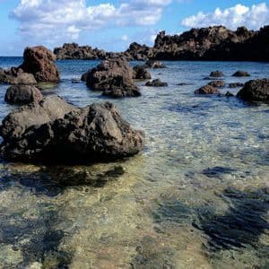 Rocce vulcaniche scure emergono da acque limpide e poco profonde lungo una costa sotto un cielo parzialmente nuvoloso.