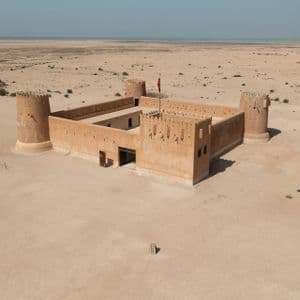 Una ripresa aerea di una fortezza quadrata color sabbia con quattro torri angolari, situata in un vasto deserto con il mare sullo sfondo.