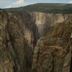 Imponenti e scure pareti rocciose di un profondo canyon, con una mesa dalla cima verde sullo sfondo e un cielo azzurro.