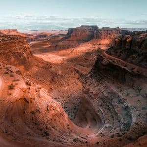 Una strada sterrata tortuosa attraversa un vasto canyon di roccia rossa sotto un cielo azzurro pallido.