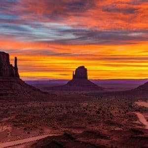 Tre grandi butte di arenaria si ergono in una valle desertica sotto un cielo striato di arancione, giallo e rosso dal tramonto.