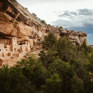 Antiche dimore in pietra scavate nella parete rocciosa, circondate da alberi verdi sotto un cielo nuvoloso.