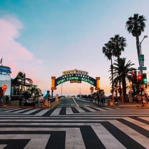L'insegna luminosa dell'ingresso del molo di Santa Monica si erge ad arco sulla strada al tramonto, con palme e un attraversamento pedonale in primo piano.